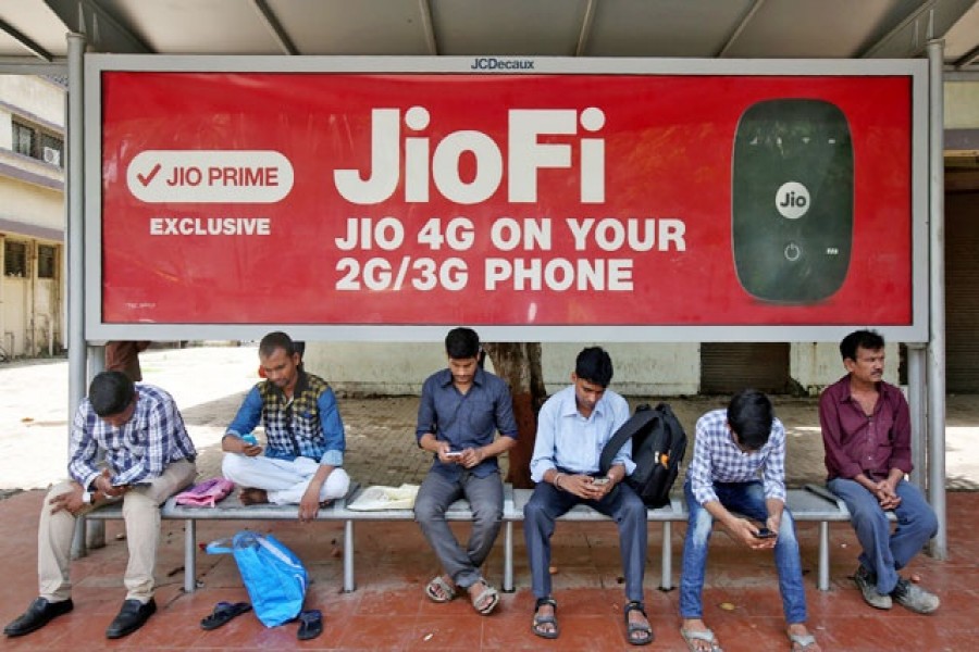 Commuters use their mobile phones as they wait at a bus stop with an advertisement of Reliance Industries' Jio telecoms unit, in Mumbai, India Jul 10, 2017. REUTERS