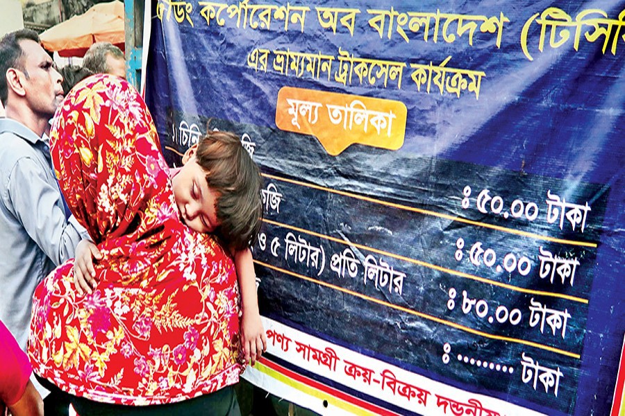 With her child falling asleep on her shoulder, a woman waits at a distance from the crowd to buy essential items from a TCB-operated truck in Topkhana area of the city on Sunday. The price hike of essentials has added to the woes of low-income people already reeling from the fallout of the coronavirus pandemic — FE photo by Shafiqul Alam